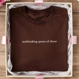 Puede incluir: Una camiseta marrón con el texto "multitasking queen of chaos" en blanco, presentada en una caja de regalo blanca con cinta rosa. La camiseta está cuidadosamente doblada, mostrando el diseño de cuello redondo.