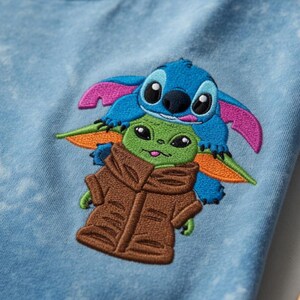 May include: Embroidered design of Stitch and Grogu (Baby Yoda) on light blue fabric. Stitch is blue with pink ears, Grogu is green with a brown coat. The design is centered on the fabric.