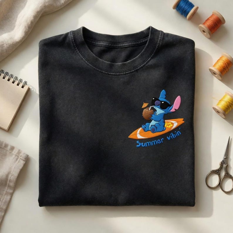 Op de afbeelding: Zwart T-shirt met een geborduurd ontwerp van een blauw stripfiguur met een zonnebril en een kokosdrankje terwijl hij surft op een surfplank. De tekst "Summer vibin'" staat eronder.