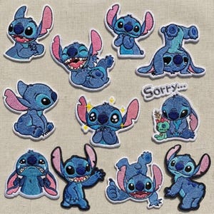 Peut inclure: Un ensemble de onze écussons brodés représentant le personnage extraterrestre bleu Stitch dans diverses poses et expressions, incluant un écusson avec le texte Sorry, ainsi qu'une petite poupée verte, tous avec des oreilles roses et des détails bleu foncé.