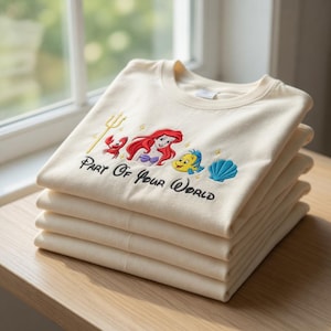 Puede incluir: Una pila de camisetas color crema con un dise&ntilde;o bordado de una sirena pelirroja, un cangrejo, un pez amarillo y una concha azul. El texto "Part Of Your World" est&aacute; bordado debajo. Las camisetas est&aacute;n cuidadosamente dobladas sobre una superficie de madera.