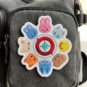 Peut inclure: Un sac à dos gris foncé avec un écusson brodé coloré. L'écusson présente des visages d'animaux de dessins animés dans un motif circulaire autour d'une boussole rouge et bleue. Le texte "STRAY KIDS" est visible sur l'écusson.