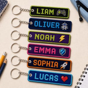 May include: Six rectangular embroidered keychains with metal rings and chains. Each features a pixelated name and icon: Liam with a game controller, Oliver with a robot, Noah with a lightning bolt, Emma with a shield, Sophia with a rocket, and Lucas with a heart.