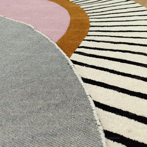 Pu&ograve; includere: Primo piano di un tappeto moderno con un design geometrico. Il tappeto presenta una combinazione di colori, tra cui grigio, rosa, marrone e bianco. Il tappeto ha strisce nere e una forma curva.