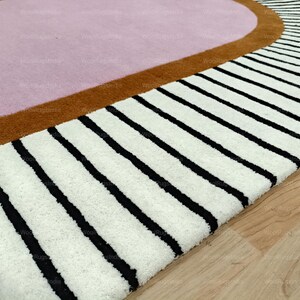 Pu&ograve; includere: Primo piano di un tappeto moderno con un design geometrico. Il tappeto presenta un centro rosa chiaro, bordato da una fascia marrone e una sezione con spesse strisce nere su sfondo bianco. Il tappeto &egrave; su un pavimento in legno.