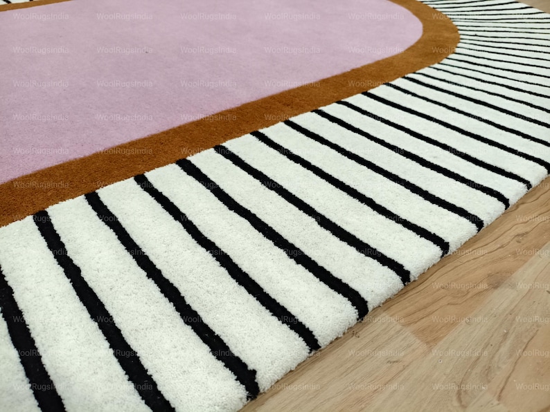 Pu&ograve; includere: Primo piano di un tappeto moderno con un design geometrico. Il tappeto presenta un centro rosa chiaro, bordato da una fascia marrone e una sezione con spesse strisce nere su sfondo bianco. Il tappeto &egrave; su un pavimento in legno.