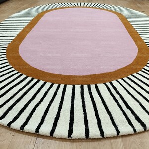 Pu&ograve; includere: Un tappeto ovale con un centro rosa, circondato da un bordo marrone e un campo bianco con strisce nere che si irradiano verso l'esterno. Il tappeto &egrave; progettato per essere posizionato su un pavimento in legno.