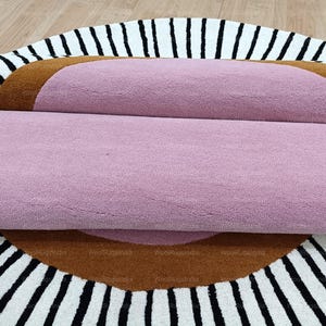 Pu&ograve; includere: Tappeto arrotolato con un centro rosa, un bordo marrone e un motivo a strisce bianche e nere. Il tappeto &egrave; realizzato in un materiale morbido e strutturato, ideale per l'arredamento della casa e misura circa 150 cm di diametro.