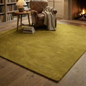 May include: A large, olive-green area rug dominates the foreground, with a soft, textured appearance. A leather armchair with a knitted throw sits to the right, near a fireplace. A small wooden side table with a lamp and books is to the left, creating a cozy living space.