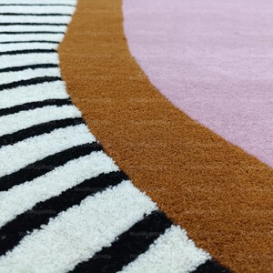 Pu&ograve; includere: Primo piano di un tappeto moderno con un design geometrico. Il tappeto presenta una sezione a strisce bianche e nere, un bordo curvo marrone e una sezione rosa. Il tappeto ha un aspetto morbido e strutturato.