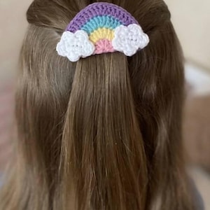 Op de afbeelding: Een gehaakte regenboog haarclip met witte wolkenaccenten. De regenboog heeft paarse, blauwe, gele en roze strepen. Het haaraccessoire is bevestigd aan bruin haar.