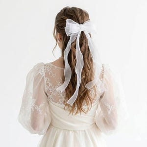 Könnte beinhalten: Eine weiße Organza-Schleife mit langen, fließenden Bändern ist im Haar befestigt. Die Schleife ist Teil einer Brautfrisur, kombiniert mit einem weißen Kleid mit Spitzendetails und transparenten Ärmeln. Das Bild ist von hinten aufgenommen.