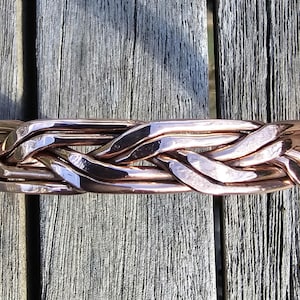 May include: A copper-colored bracelet with a woven design. The bracelet is made of multiple strands of metal that are intertwined to create a braided pattern. The bracelet is shown on a wooden surface.
