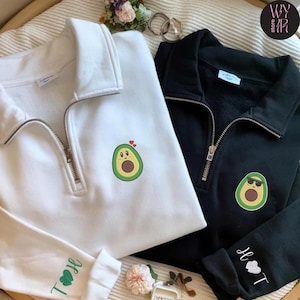 Könnte beinhalten: Zwei Sweatshirts mit Viertelreißverschluss, eines in Weiß und eines in Schwarz. Jedes hat ein gesticktes Avocado-Design. Das weiße Sweatshirt hat eine überraschte Avocado mit Herzen, das schwarze eine Avocado mit Sonnenbrille. Ärmel mit gestickten Initialen.