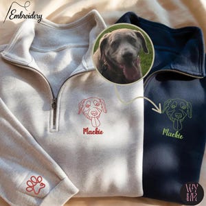 May include: Two quarter-zip sweatshirts, one light gray and one navy blue, featuring custom dog embroidery. The light gray sweatshirt has a red paw print on the sleeve and a red dog face with the name "Mackie". The navy blue sweatshirt has a green dog face with the name "Mackie".