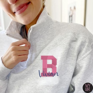 Puede incluir: Sudadera gris claro con cremallera de cuarto de cuello, con una gran letra "B" rosa y blanca y el nombre "Vivian" bordado en la parte delantera. La sudadera tiene un cuello alto y una cremallera plateada.