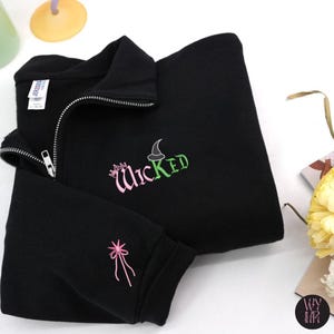 May include: Black zip-up sweatshirt with the word "Wicked" embroidered in pink and green. A pink bow is embroidered on the sleeve. The sweatshirt has a silver zipper and a high collar.