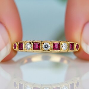 May include: A gold ring with alternating square-cut red gemstones and round white diamonds. The ring has a delicate, vintage-inspired design with a repeating pattern of gemstones and diamonds. The ring is held up, showcasing its intricate details.