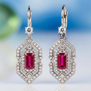 May include: A pair of Art Deco-style earrings with a rectangular, deep red gemstone center. The earrings feature a geometric design with small, clear gemstones and a lever-back closure. The metal appears to be silver or white gold.