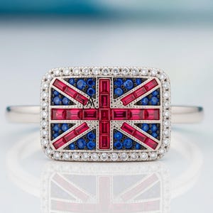 May include: A silver ring featuring a rectangular design of the Union Jack, the flag of the United Kingdom. The flag design is made of red, blue, and white gemstones. The ring is set with a halo of small white gemstones.