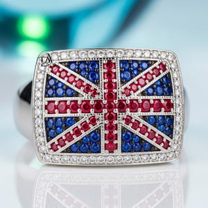May include: A silver ring with a rectangular face featuring a Union Jack design. The flag is rendered with blue and red gemstones, surrounded by a border of clear stones. The ring is set against a reflective surface.