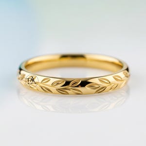Puede incluir: Un anillo dorado con un patrón de hojas grabado alrededor de la banda. El anillo tiene un acabado pulido y un diseño de banda simple. El anillo probablemente está hecho de oro o un metal dorado. Una joya.
