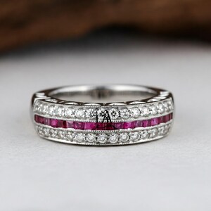 Peut inclure: Une bague en argent ornée de deux rangées de rubis carrés et d'une rangée de petits diamants ronds. La bague présente un motif complexe sur les bords et les pierres précieuses sont serties dans un sertissage à griffes. La bague est sur une surface blanche.