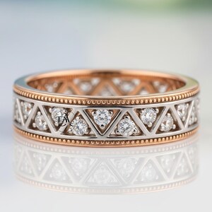 May include: A two-tone ring with a rose gold interior and a silver exterior. The silver band features a repeating triangular pattern with small, round diamonds. The top and bottom edges are lined with small, gold beads. The ring is displayed on a reflective surface.