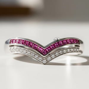 May include: A silver V-shaped ring with a row of square-cut pink gemstones above a row of small, round, clear gemstones. The ring is set on a white surface, with soft shadows.