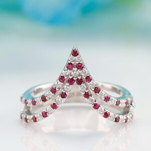 Op de afbeelding: Een zilveren ring met een dubbele band, versierd met kleine, ronde rode en witte edelstenen. De ring heeft een centrale, omhoog wijzende V-vorm, eveneens versierd met rode edelstenen. De edelstenen zijn in pavé-stijl gezet.