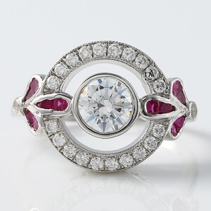 Op de afbeelding: Een zilveren ring met een grote, ronde, heldere edelsteen in het midden, omringd door kleinere heldere stenen. De ring heeft rode edelstenen aan elke kant, in een bloemmotief. Op een witte achtergrond.