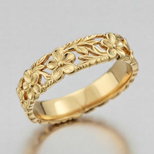 Pode incluir: Um anel dourado com um design floral. O anel apresenta entalhes detalhados de flores e folhas, criando um padrão intrincado ao redor da banda. O anel é mostrado em um fundo claro.