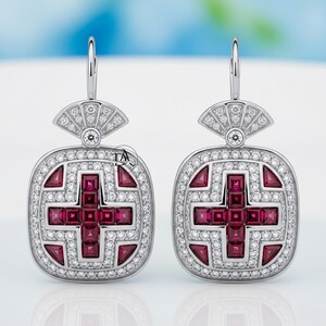 Peut inclure: Une paire de boucles d'oreilles en argent avec un motif carré, représentant une croix rouge et entourée de petites pierres précieuses transparentes. Le haut de chaque boucle d'oreille présente un motif en forme d'éventail avec d'autres pierres précieuses.