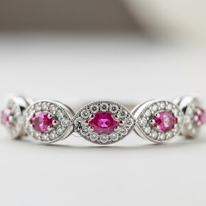 May include: A silver ring featuring a series of oval-shaped, bright pink gemstones, each surrounded by a halo of small, clear stones. The ring has a delicate, vintage-inspired design.