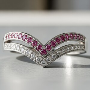 May include: A V-shaped ring with two bands. The top band is lined with small, pink gemstones, while the bottom band is lined with small, clear gemstones. The ring is made of a silver-colored metal.