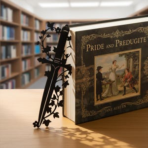 Könnte beinhalten: Ein schwarzer Metall-Lesezeichen in Schwertform mit floralen Verzierungen, neben einem Hardcover-Buch platziert. Der Buchumschlag zeigt den Titel "Pride and Prejudice" und eine Illustration aus der Zeit. Das Lesezeichen liegt auf einer Holzoberfläche.