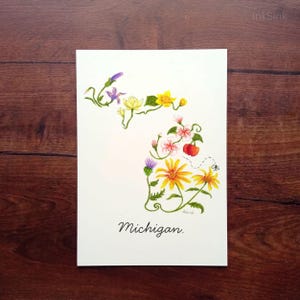 May include: A white rectangular print featuring a botanical illustration of the state of Michigan outlined by various colorful flowers, green stems, and a small red cherry. The word Michigan is printed in black cursive script at the bottom.