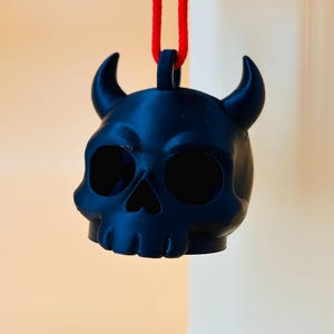 May include: A black skull-shaped pendant with curved horns, suspended by a red cord. The pendant has large eye sockets and a nose cavity. The text "PERFUME COM" is visible at the top. The pendant is likely a decorative item.