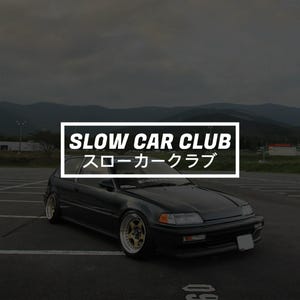 May include: A dark-coloured car with gold-coloured alloy wheels is parked on an asphalt surface. The car has the words "SLOW CAR CLUB" and Japanese characters in a white box. Mountains and a cloudy sky are in the background.