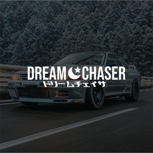 May include: A gray sports car with the text "DREAM CHASER" in white, along with Japanese characters and a crescent moon and star symbol. The car is on a road with a blurred forest background.