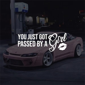 May include: A pink sports car with white rims and the text "YOU JUST GOT PASSED BY A Girl" in white. A white lip print and a heart are also on the car. The car is parked at a petrol station at night.