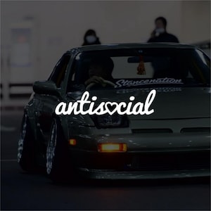 Puede incluir: Un coche deportivo verde oscuro con la palabra "antisocial" en escritura blanca en la parte delantera. El coche tiene una suspensión rebajada y llantas personalizadas. El texto incluye un símbolo de corazón. El coche está aparcado en una zona poco iluminada.