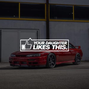 May include: A red sports car with dark grey wheels and a rear spoiler. A white rectangular graphic overlay features a thumbs-up icon and the text "YOUR DAUGHTER LIKES THIS." The car is parked on a grey surface in front of a building with windows.