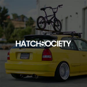 May include: A yellow Honda Civic with a black BMX bike on a roof rack. The white text "HATCHSOCIETY" is across the rear window. The car has custom wheels and a rear spoiler. The car is a hatchback.