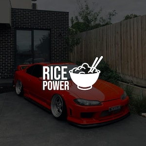 Peut inclure: Une voiture de sport rouge avec le texte "RICE POWER" en blanc. Un bol blanc de nourriture avec des baguettes est sur le côté droit de la voiture. La voiture est garée devant une maison.