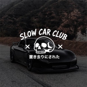 May include: A black sports car featuring a white graphic overlay with the text SLOW CAR CLUB and Japanese characters, centred above a stylised skull icon with cross symbols on either side, positioned on a dark asphalt road surface.