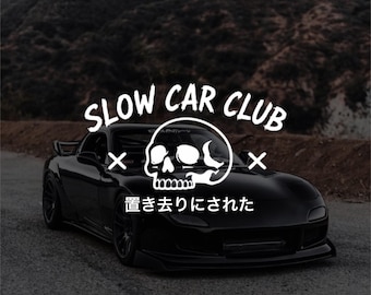 Könnte beinhalten: Ein schwarzer Sportwagen auf einer Straße mit einer weißen Grafiküberlagerung, die einen Totenkopf, den Text SLOW CAR CLUB und den japanischen Text 置き去りにされた zeigt, was zurückgelassen oder verlassen bedeutet.