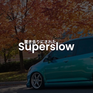 May include: A teal-colored car with the word "superslow" in white text. The car is parked on a road with a backdrop of trees with orange and yellow leaves. The Japanese text above the word "superslow" is also visible.