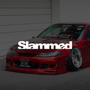 Puede incluir: Un coche deportivo rojo con la palabra "Slammed" en blanco en el capó. El coche tiene llantas personalizadas y un perfil bajo, con una matrícula que dice "326". El coche está estacionado sobre una superficie gris.