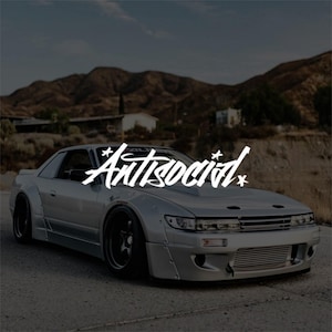 Pode incluir: Um carro desportivo prateado com rodas pretas e um kit de carroçaria personalizado. A palavra "Antisocial" está escrita em branco no para-brisas. O fundo apresenta uma cordilheira e um céu nublado.